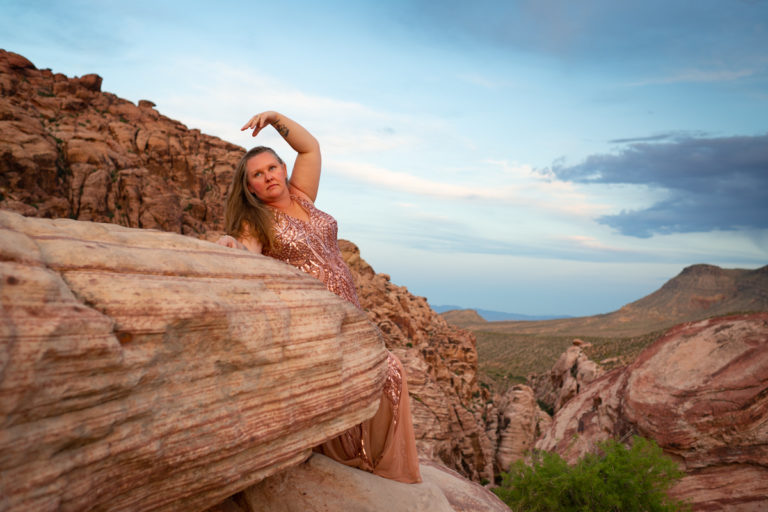 A plus-size white woman in a pink sequined gown with long dark blonde hair arches an arm and looks off to the distance while leaning on a boulder in a desert landscape.