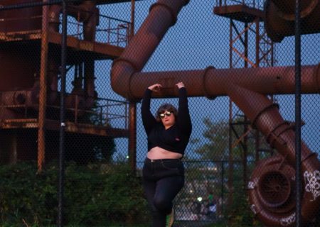 A fat white woman with shoulder-length brown hair leans and hangs onto a chain-link fence with big industrial pipes behind her. She's wearing a long-sleeved crop top, jeans and sunglasses.