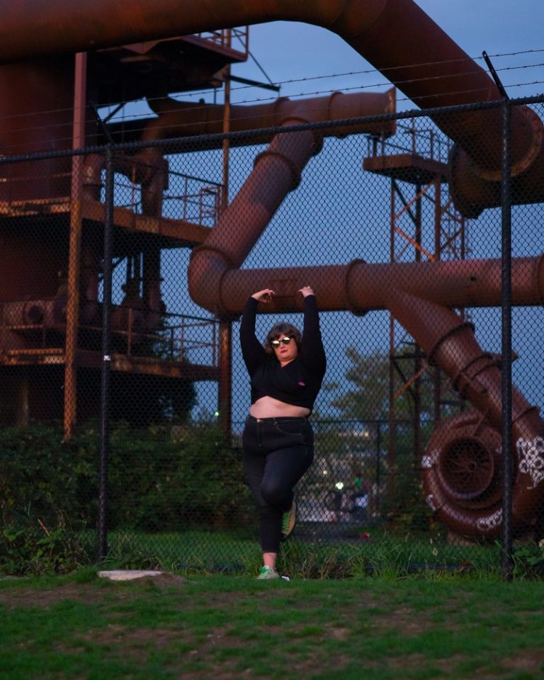 A fat white woman with shoulder-length brown hair leans and hangs onto a chain-link fence with big industrial pipes behind her. She's wearing a long-sleeved crop top, jeans and sunglasses.