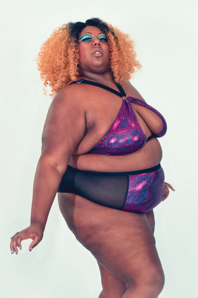A Black model with red hair and a purple underwear set on looks confidently at the camera
