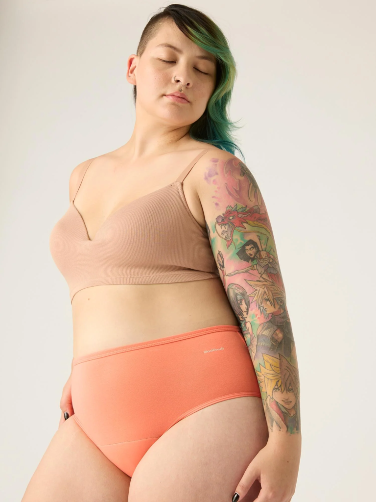 A woman with green side shaved hair wearing a beige bra and orange panties