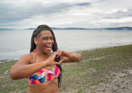 A Black trans woman in a colorful bikini smiles on a seaweedy beach and makes a heart shape with her hands.