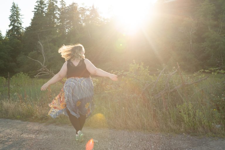 Lindley, a fat white woman, twirls with her hair and skirt flying out on a gravel road with the sun shining through trees behind her.