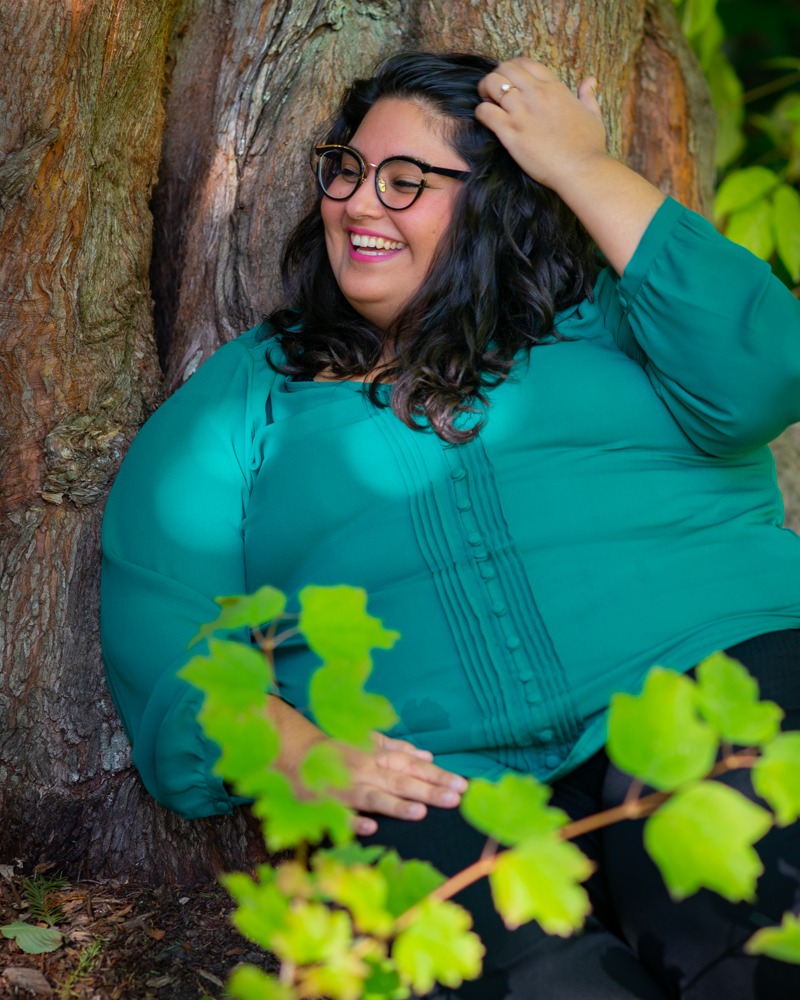 A fat woman of Iranian descent, with long black hair, glasses and a teal blouse, relaxes against a tree trunk with one hand in her hair.