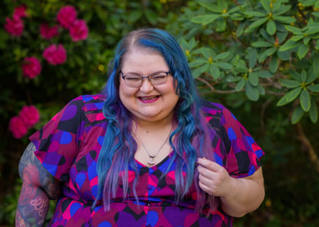 A superfat white woman with long blue and purple hair, glasses and arm tattoos laughs hard in a garden with bright pink flowers. She's wearing a Mjolnir necklace and a colorful top that coordinates with the flowers.