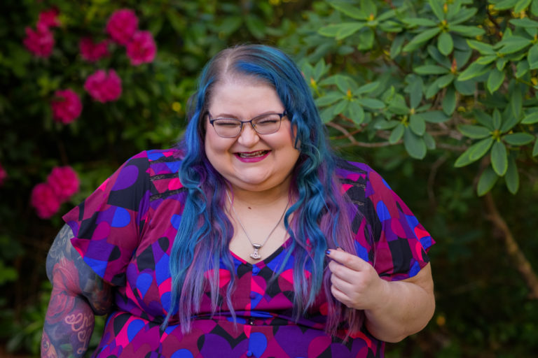 A superfat white woman with long blue and purple hair, glasses and arm tattoos laughs hard in a garden with bright pink flowers. She's wearing a Mjolnir necklace and a colorful top that coordinates with the flowers.