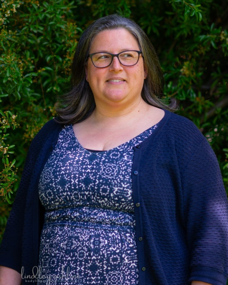 A plus-size white woman with mid-length gray hair in a blue dress and cardigan, standing in a garden, looking away from the camera and smiling.