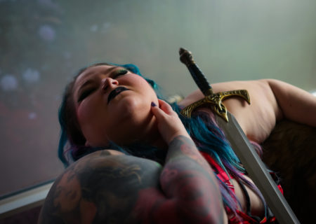 A superfat woman with arm tattoos and blue hair leans back in a chair with her eyes closed, one hand on her jawline and a fantasy sword propped against her in low lighting.