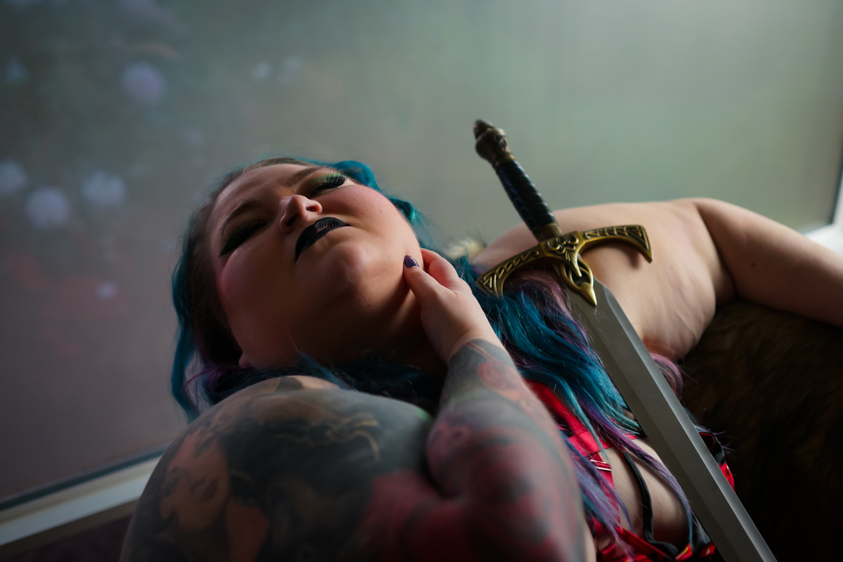 A superfat woman with arm tattoos and blue hair leans back in a chair with her eyes closed, one hand on her jawline and a fantasy sword propped against her in low lighting.