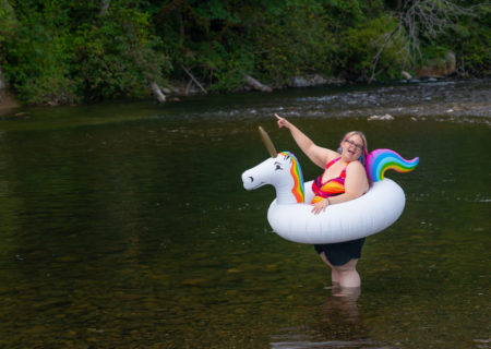 A fat white woman with pink hair and a colorful bathing suit stands in a shallow river, laughing and pointing at something in the distance, while wearing an inflatable unicorn inner tube-style float.