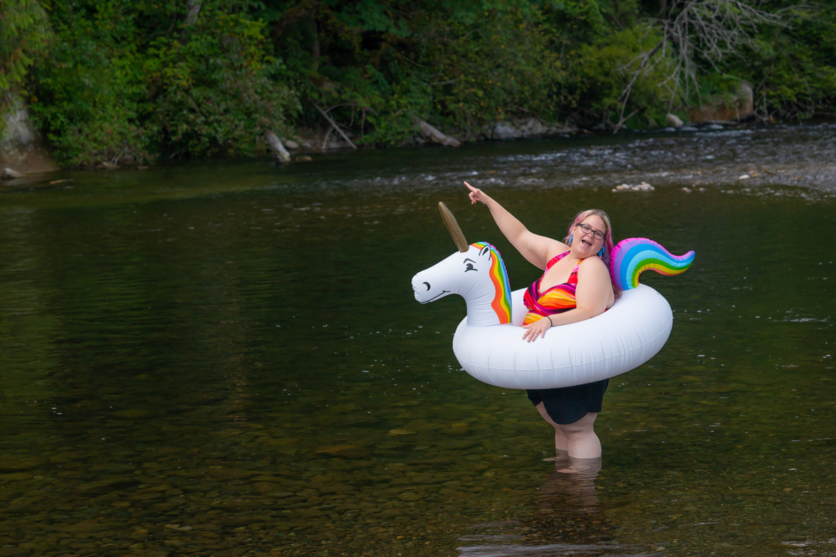 A fat white woman with pink hair and a colorful bathing suit stands in a shallow river, laughing and pointing at something in the distance, while wearing an inflatable unicorn inner tube-style float.