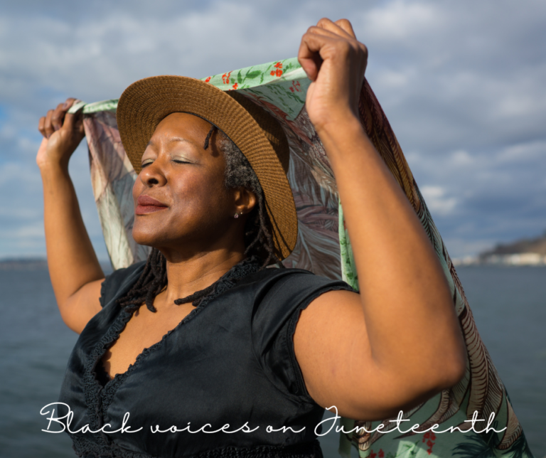 A plus-size Black woman closes her eyes and smiles in the sunshine while holding a wing-pattern scarf behind her head in front of water and a cloudy sky.