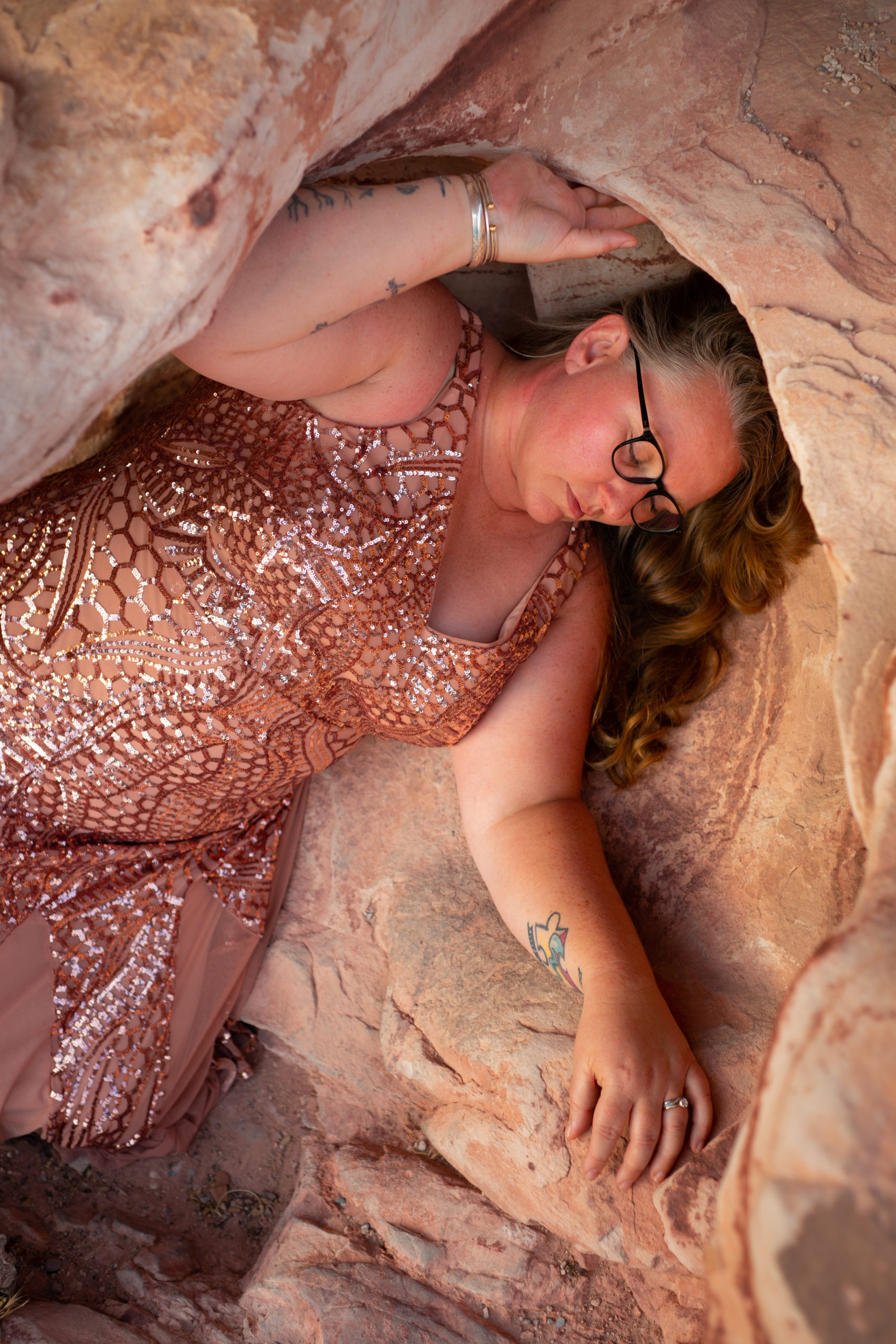 A plus-size woman in a sparkly pink dress, nestled in pink rocks in the desert at a body-positive portrait session.