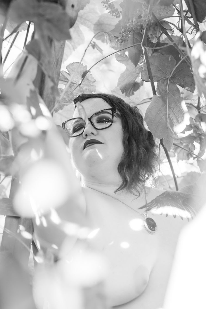A plus-size nonbinary person looks down at the camera during a fat-positive outdoor boudoir session in a black and white image.
