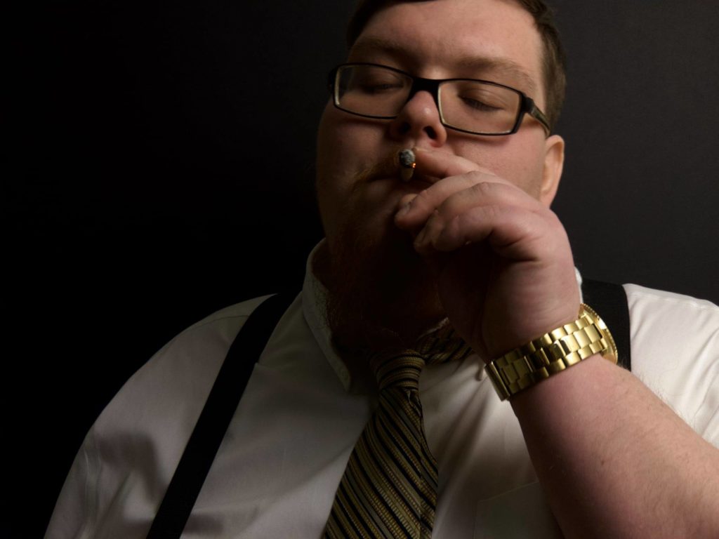 A fat white man wearing a white shirt with suspenders, black framed glasses and a gold watch smokes a cigar