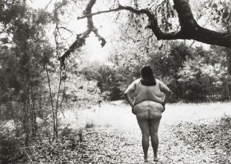 A black-and-white photograph of a fat feminine figure standing nude in a forest clearing, under a huge tree branch. Their hands are on their hips.