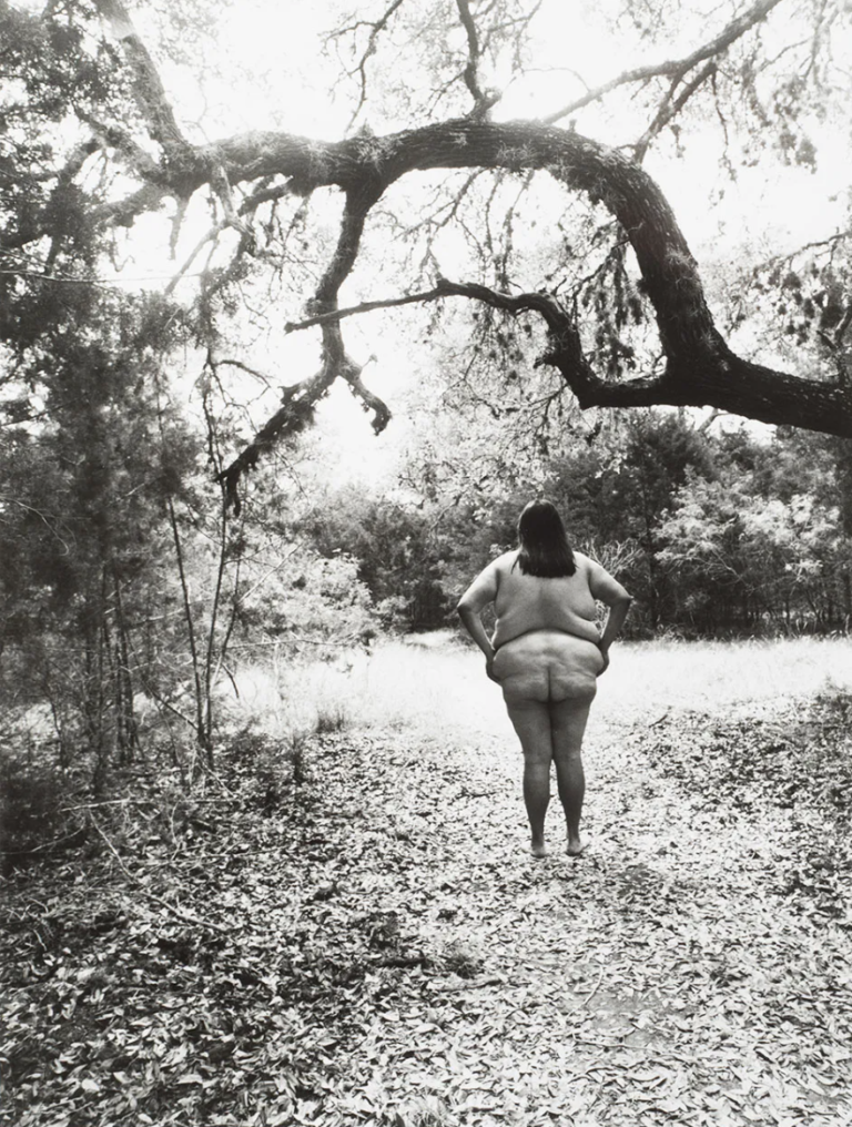 A black-and-white photograph of a fat feminine figure standing nude in a forest clearing, under a huge tree branch. Their hands are on their hips.