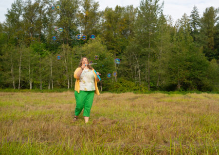 A fat white woman in capris, a top and cardigan blows bubbles in a field of grass during a body-positive portrait shoot in Washington state.