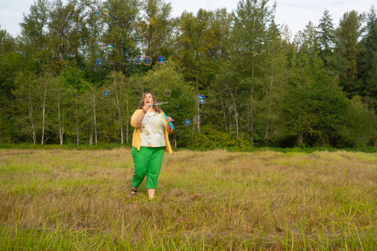 A fat white woman in capris, a top and cardigan blows bubbles in a field of grass during a body-positive portrait shoot in Washington state.