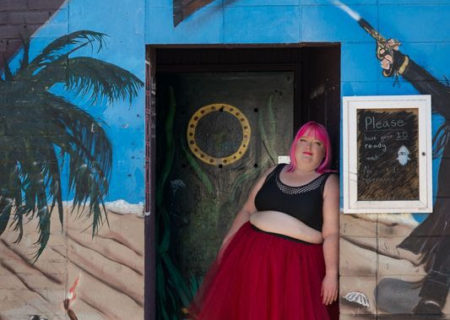 A plus-size femme person with bright pink hair, a tutu skirt and crop top leans against the doorway of a building painted in a pirate theme.