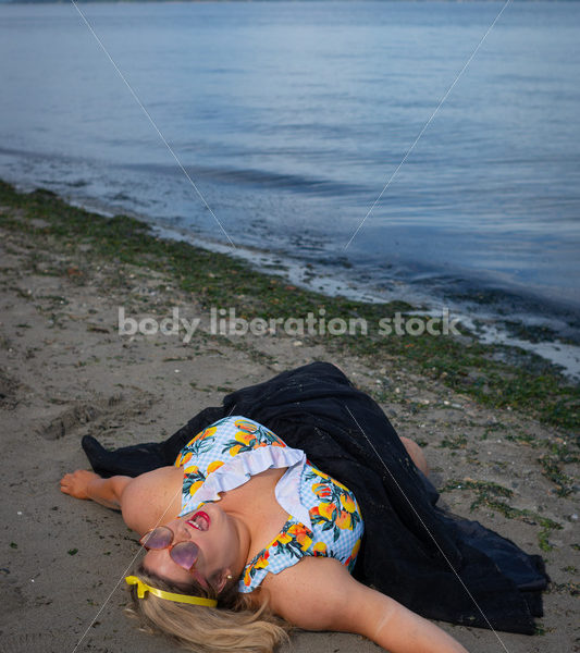 Body-Positive Stock Photo: Plus Size Woman in a Blue Swimsuit and Skirt Coverup Lays on the Sand Calling Out to the Sky - Body Liberation Photos & Stock