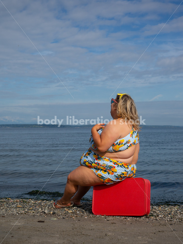 Body-Positive Stock Photo: Plus Size Woman in a Swimsuit Sits on a Red Suitcase in Front of the Ocean - It's time you were seen ⟡ Body Liberation Photos