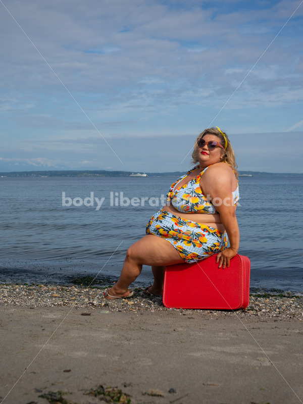 Body-Positive Stock Photo: Plus Size Woman in a Swimsuit Sits on a Red Suitcase in Front of the Ocean with an Alluring Look - It's time you were seen ⟡ Body Liberation Photos