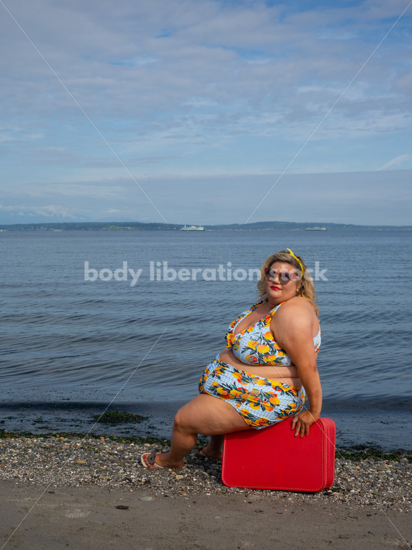 Body-Positive Stock Photo: Plus Size Woman in a Swimsuit Sits on a Red Suitcase in Front of the Ocean with an Alluring Look - It's time you were seen ⟡ Body Liberation Photos