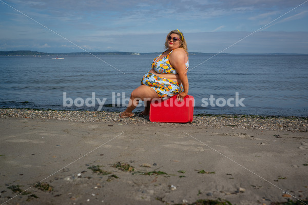 {Fat Glam Summer} Body-Positive Stock Photo: Plus Size Woman in a Swimsuit Sits on a Red Suitcase in Front of the Ocean with an Alluring Look - Body Liberation Photos & Stock