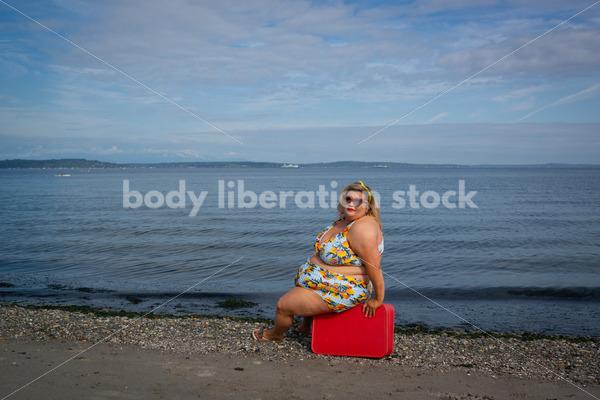 {Fat Glam Summer} Body-Positive Stock Photo: Plus Size Woman in a Swimsuit Sits on a Red Suitcase in Front of the Ocean with an Alluring Look - Body Liberation Photos & Stock