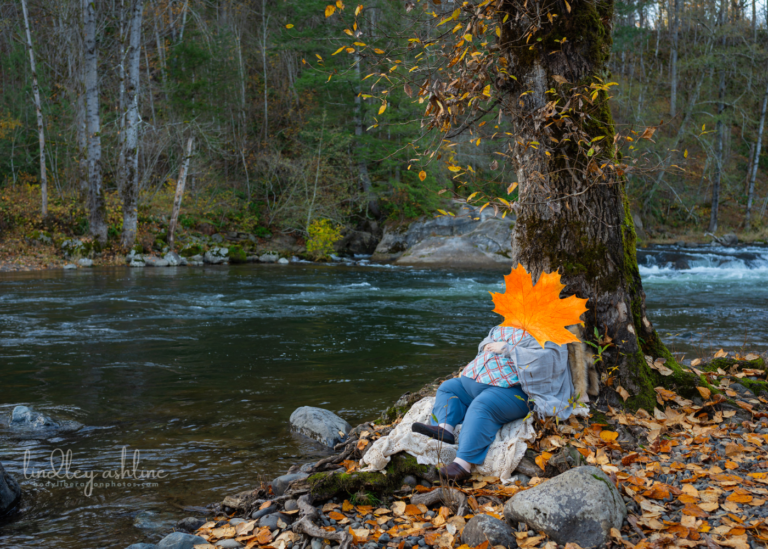 A fat pregnant white woman leans against a tree with yellow leaves beside a river. Her face is covered with a maple leaf sticker for privacy.