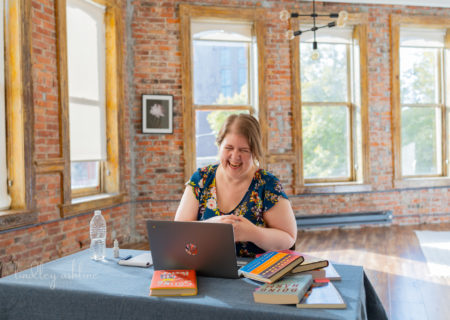 A fat white woman laughs in front of a laptop at a table in a large, brightly-lit room with brick walls and big windows. Books are scattered on the table around the laptop. Part of a fat-positive small business branding session in Seattle, WA