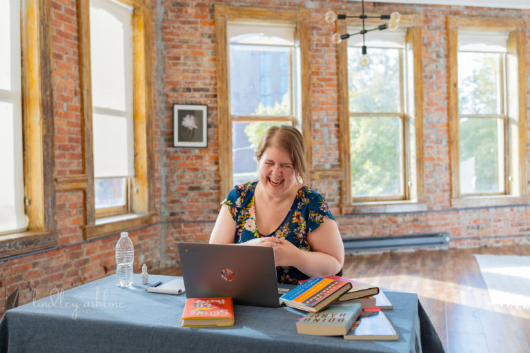 A fat white woman laughs in front of a laptop at a table in a large, brightly-lit room with brick walls and big windows. Books are scattered on the table around the laptop. Part of a fat-positive small business branding session in Seattle, WA