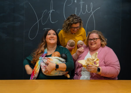 Three women laugh hard in front of a chalkboard with Jet City Doulas written on it. One is holding a model of the pelvic bones, one is holding a realistic baby doll in a sling and one is holding two different-colored knitted model breasts to her chest.