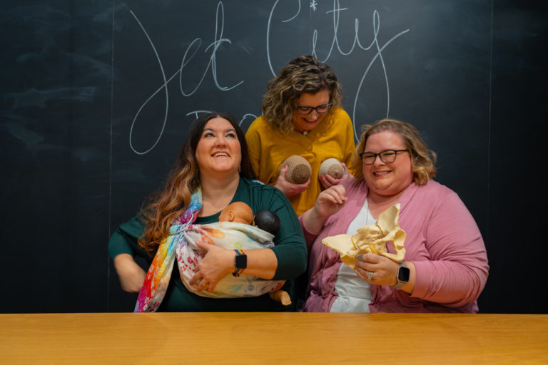 Three women laugh hard in front of a chalkboard with Jet City Doulas written on it. One is holding a model of the pelvic bones, one is holding a realistic baby doll in a sling and one is holding two different-colored knitted model breasts to her chest.