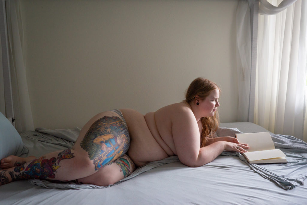 A photograph of the same feminine person lying down and reading, with tattoos on their legs (but not the mermaid tattoo from the painting).