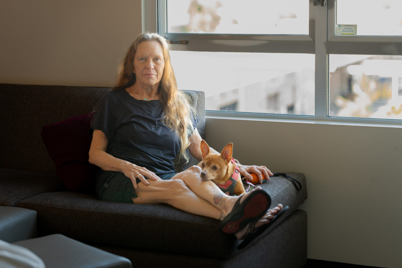 A thin, older white woman with wrinkled skin sitting solemnly on a hotel sofa by a window with her dog.