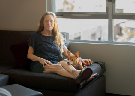 A thin, older white woman with wrinkled skin sitting solemnly on a hotel sofa by a window with her dog.