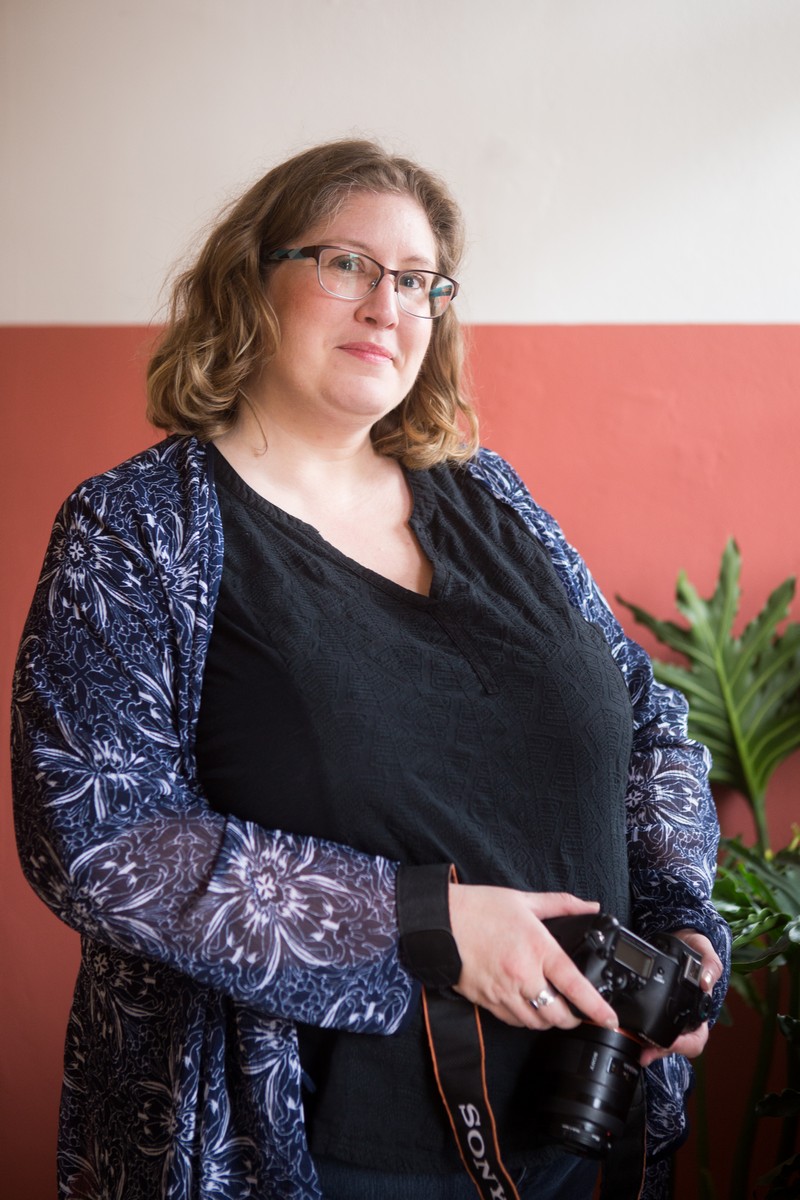 Lindley, a fat white woman wearing glasses and a casual outfit, holds a camera and smiles at the camera in window light.