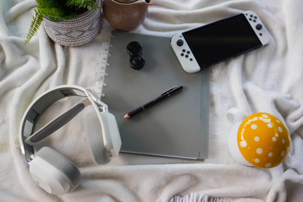 A flat lay of some of Kyra Sutherland's ADHD and autism coaching tools, including a notebook, potted plant, Switch handheld game system, coffee mug, headphones, pen and earplugs.