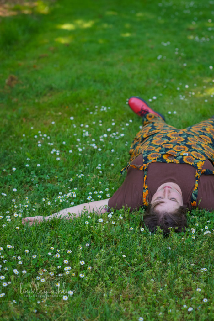 A non-binary person with short hair and casual clothes lies on their back in the grass and gazes up at the sky during a queer-friendly Seattle small business branding photo session for coaches.