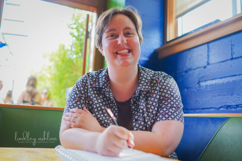A non-binary person with short hair and casual clothes smiles and takes notes at a restaurant table during a Seattle small business branding photo session for coaches.