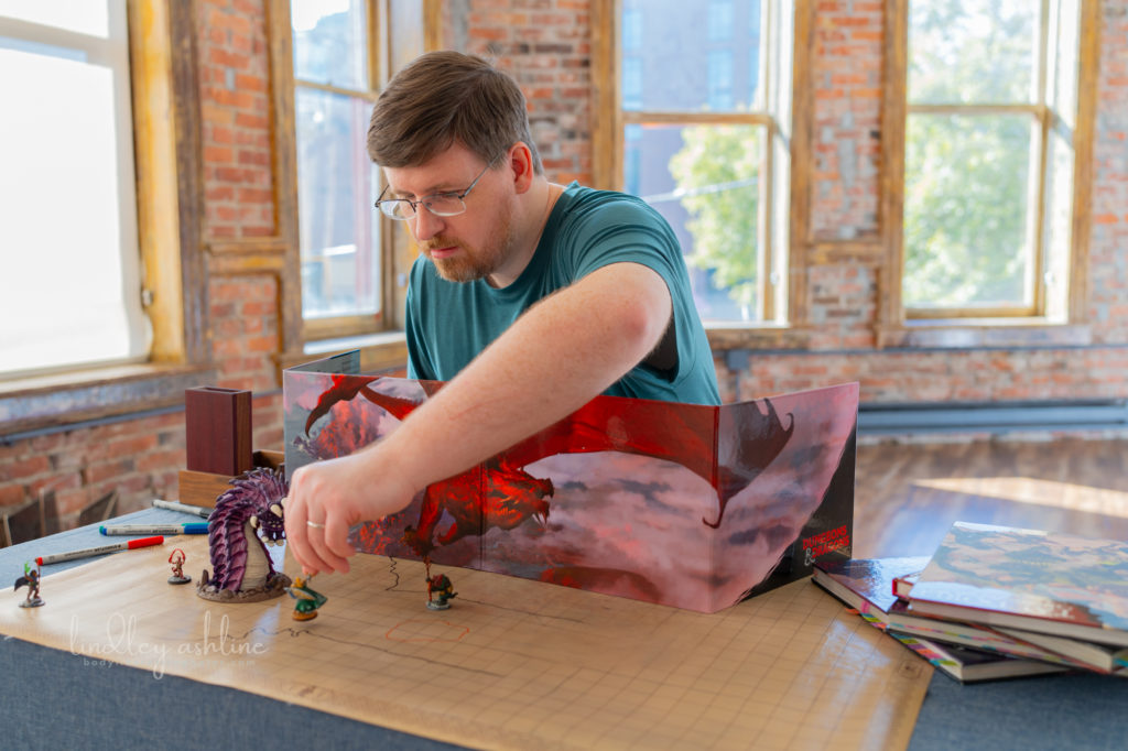 A mid-size white male gamer with a D&amp;D DM screen setup prepares to roll dice in a sunlit room at a Seattle professional portrait photo session.