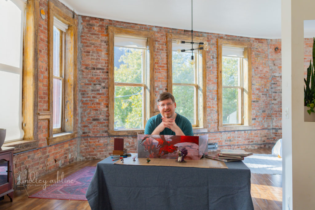 A mid-size white male gamer with a D&amp;D DM screen setup prepares to roll dice in a sunlit room at a Seattle professional portrait photo session.