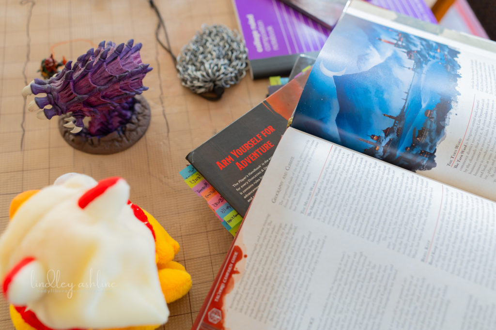 A variety of gamer and geek accessories laid out on top of a dry erase D&D map, including D&D books, dice pouch, mini figurine, and a chocobo plushy.