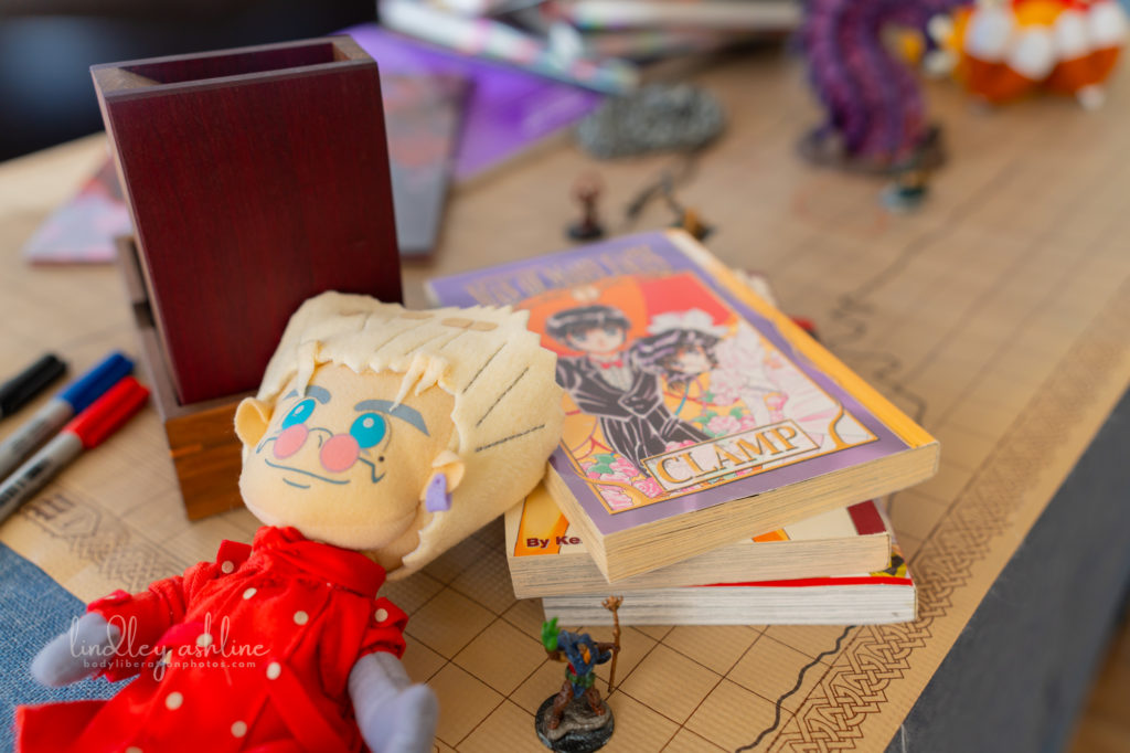 A variety of gamer, geek and anime accessories laid out on top of a dry erase D&D map, including a dice tower, anime plushy and manga volumes.