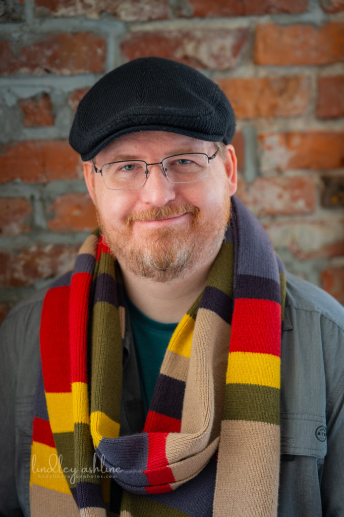 Photo of Karl Reichert, LMHC, wearing the 4th doctor&#039;s scarf (from Doctor Who), glasses, and a black driving cap. Karl is a white man with reddish blond beard.