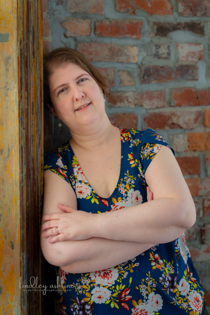 A plus-size white woman in a sleeveless top leans against a wood window frame in front of a brick wall, at a Seattle body-positive business branding session.