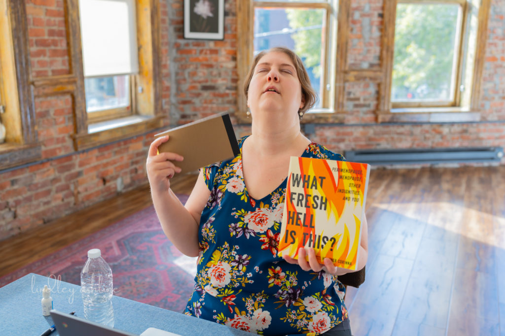 A plus-size menopausal white woman in a sleeveless top fans herself with a notebook and a copy of the book What Fresh Hell is This? at a Seattle body-positive business branding session.