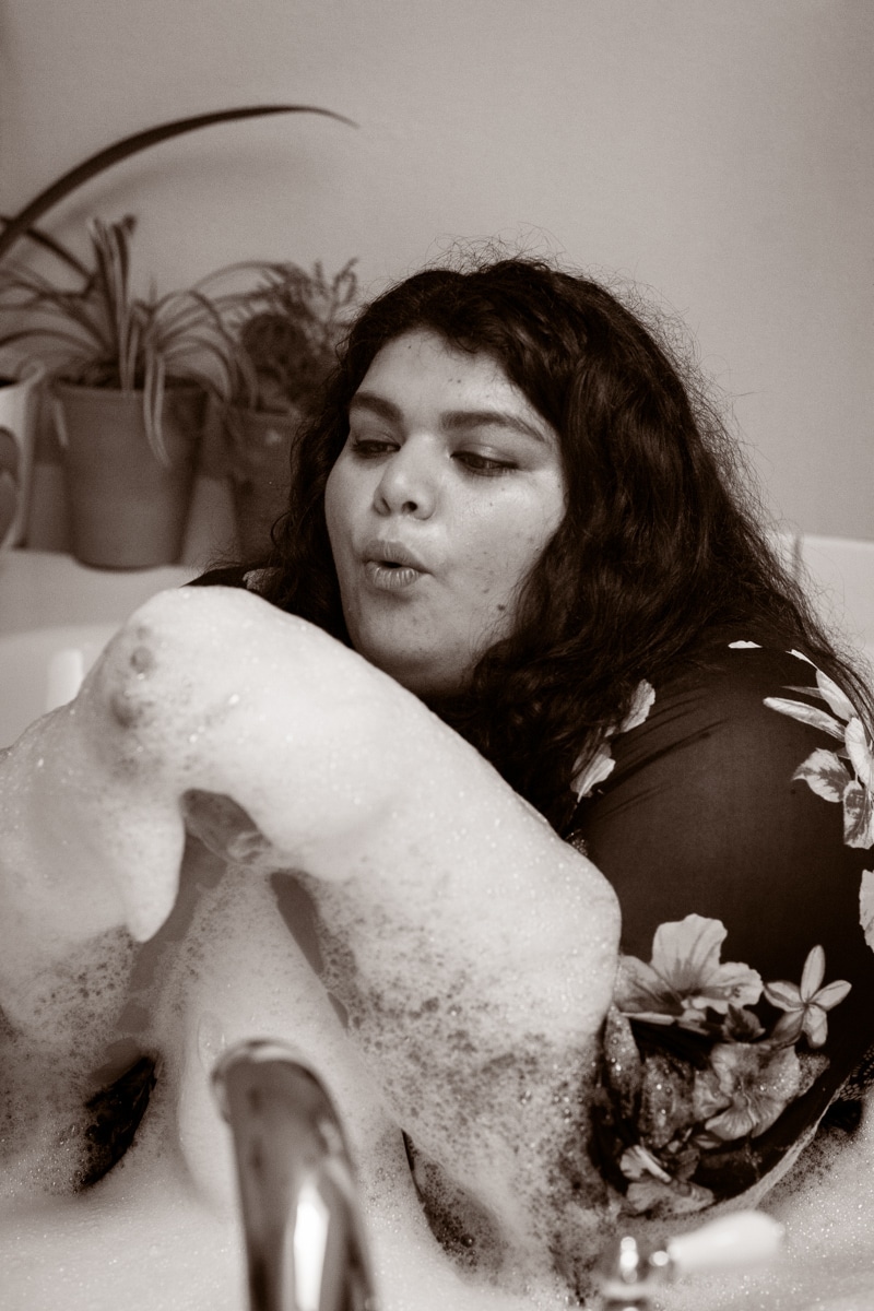 A brown-toned photo of a woman with black hair and brown skin blowing on bubbles in a bubble bath at a fat-positive Seattle boudoir shoot.